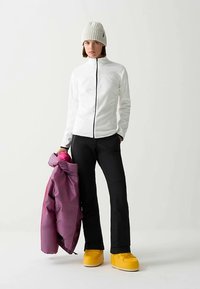 Fehér cipzáros dzseki, fekete nadrág és élénksárga lábbeli. Lila dzsekit tart a kezében. A texturált kötött sapka kiegészíti a ruházatot.