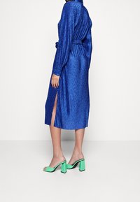 Robe en satin bleu avec des rayures verticales texturées, manches longues, fentes latérales et taille cintrée. Associée à des sandales à talons bloc vert.
