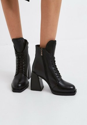 Black leather lace-up ankle boots with block heels and side zippers worn on bare legs against a white background.