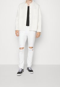 Man med en krämfärgad skjorta, svart tröja, trasiga vita jeans och svart-vita sneakers som står mot en enkel vit bakgrund.