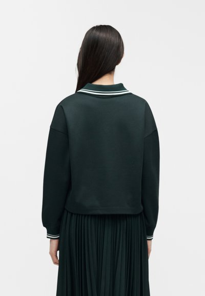 Femme aux longs cheveux foncés portant un haut à manches longues vert foncé avec un col et des poignets rayés blancs, ainsi qu'une jupe plissée assortie, vue de dos.