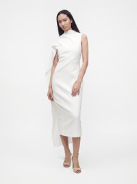 Femme aux longs cheveux foncés portant une robe midi blanche sans manches avec un col haut et un détail drapé asymétrique, debout devant un fond blanc.