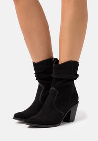 Czarne zamszowe botki za kostkę o luźnym kroju, z ostrym noskiem i słupkowym obcasem. Posiadają kontrastowe przeszycia oraz boczny zamek błyskawiczny.