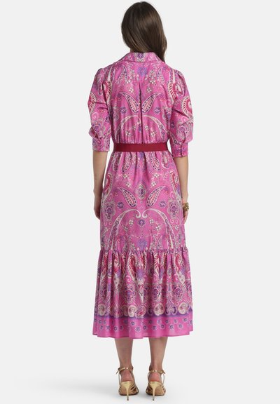 Kvinde iført en pink midikjole med blomsterprint, puffede ærmer og et rødt bælte, stående med ryggen mod en hvid baggrund.