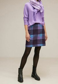 Kvinna klädd i en lila tröja och halsduk, tillsammans med en rutig kjol i lila och blå nyanser, svarta strumpbyxor och ankelskor, står avslappnat.