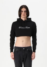 Black cropped hoodie with drawstring hood, featuring glossy "Rider Room" text in a decorative font on the front. Worn with dark jeans.