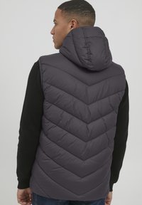 Tamno siva puffer prsluk s kapuljačom, s chevron štepanim uzorkom i prednjim džepovima, nosi se preko crne majice s dugim rukavima.