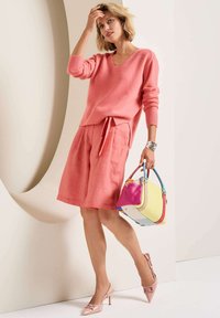 Rosa Kaschmirpullover mit V-Ausschnitt, kombiniert mit rosa Leinen-Shorts. Accessoires sind eine mehrfarbige Handtasche und rosa Absatzschuhe.