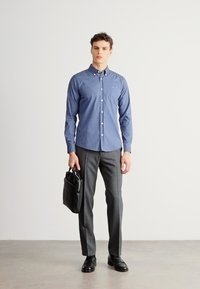 Modrá košeľa na zapínanie s jemnou textúrou, doplnená úzkymi sivými nohavicami. Model drží čiernu koženú aktovku a nosí čierne topánky.