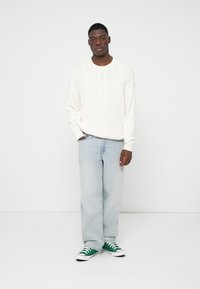 Λευκό πουλόβερ σε πλέκτυνη ύφανση με μακριά μανίκια, ανοιχτό μπλε τζιν ίσιας γραμμής και πράσινα χαμηλά αθλητικά παπούτσια. Απλά, υφασμάτινα σχέδια σε όλη την επιφάνεια.