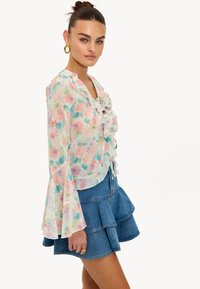 Blouse fleurie transparente avec volants, manches longues évasées et couleurs pastel en rose et vert, associée à une jupe à volants en denim.