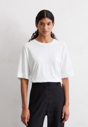 White short-sleeve t-shirt made of lightweight cotton, featuring a chest pocket. Paired with black tailored pants for a casual look.