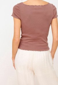 Femme portant un haut mauve ajusté à manches courtes avec un ourlet en dentelle et un pantalon en lin blanc ample, se tenant avec les mains dans les poches, dos à l'appareil photo.