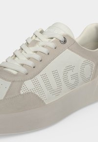Gros plan d'une basket beige et blanche avec le nom de la marque perforé sur le côté et des lacets blancs passés à travers des œillets en métal.