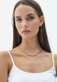 Collana in metallo dorato con un design flessibile e ondulato, indossata attorno a un collo nudo. Il modello indossa un top bianco su uno sfondo neutro.