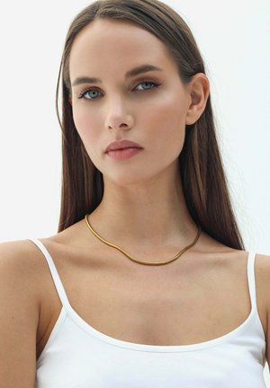 Collana in metallo dorato con un design flessibile e ondulato, indossata attorno a un collo nudo. Il modello indossa un top bianco su uno sfondo neutro.