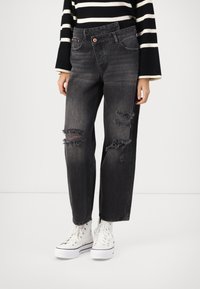 Črne jeans hlače z ohlapnim krojem, z vdrtinami in raztrganimi robovi. Ujemajo se z belimi športnimi copati z visokim zgornjim delom.
