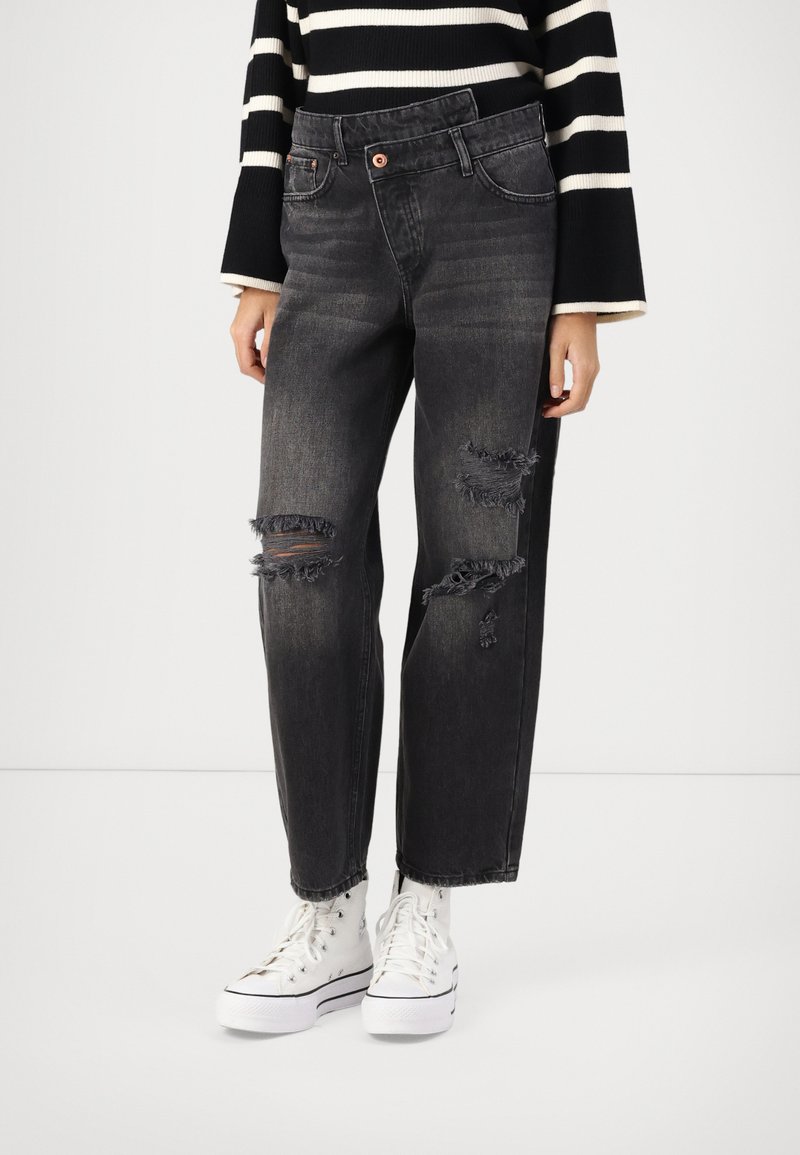 Črne jeans hlače z ohlapnim krojem, z vdrtinami in raztrganimi robovi. Ujemajo se z belimi športnimi copati z visokim zgornjim delom.