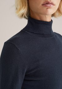 Tumši jūraszilos krāsas augsto kaklu džemperis, mīksts adījums, pieguļošs piegriezums, ar augstu apkakli un bezšuvju pleciem, nodrošinot tīru siluetu.