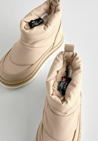 Beige geïsoleerde laarzen met een gevoerde nylon buitenkant, nepbont binnenvoering en een gestructureerde rubberen zool. Voorzien van treklabels voor gemakkelijk aan- en uittrekken.