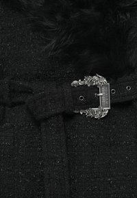 Črna teksturirana tkanina z kosmatim ovratnikom, okrašena z bogatim srebrnim zaponkom kot podrobnostjo pasu. Kovinski poudarki dodajo kontrast in zanimivost.