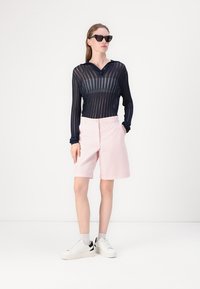 Marinblå ribbade långärmade skjorta, ljusrosa skräddarsydda shorts, vita sneakers med svarta detaljer, modell bär mörka solglasögon, framför en enfärgad bakgrund.