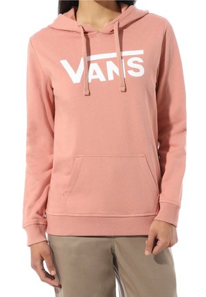 Osoba oblečená v růžové mikině Vans s přední kapsou a stahovacími šňůrkami, v kombinaci s béžovými kalhotami, stojící s uvolněnýma rukama.