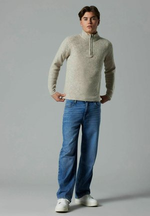 Ljusbeige zip-tröja med ribbad textur, kombinerad med ljusblå raka jeans och vita sneakers mot en neutral bakgrund.
