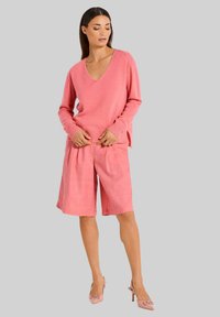 Rosa gestrickter V-Ausschnitt-Pullover kombiniert mit rosa Leinen-Shorts und hellrosa High Heels. Hat eine lockere Passform und gerippte Bündchen.