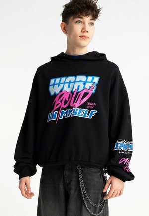 Młoda osoba z krótkimi kręconymi włosami, ubrana w czarną bluzę z kapturem z kolorowym, wyrazistym napisem oraz ciemne spodnie z srebrnym łańcuszkiem jako dodatkiem.