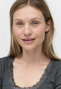 Grå top med en bølget blondeudskæring og blød tekstur; lys hudtone, glat blond hår, blå øjne og subtil makeup.