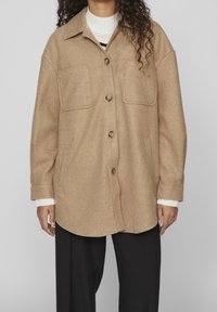 Giacca in misto lana color beige con una vestibilità rilassata, dotata di una chiusura a bottoni, due tasche sul petto e un orlo arrotondato. Indossata sopra un maglione bianco.
