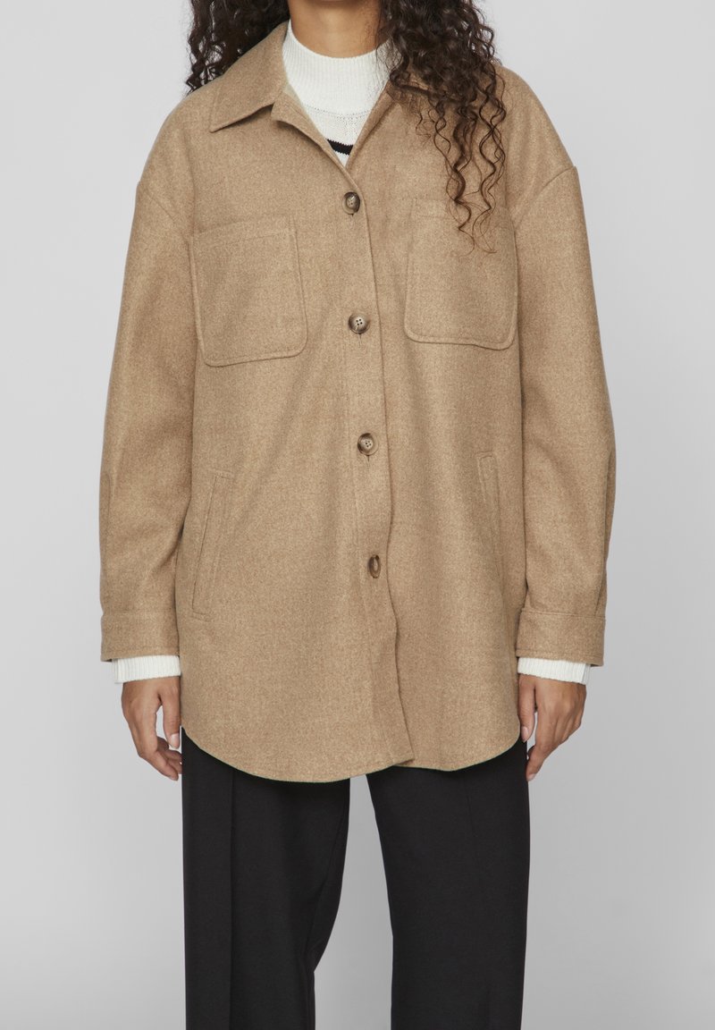 Giacca in misto lana color beige con una vestibilità rilassata, dotata di una chiusura a bottoni, due tasche sul petto e un orlo arrotondato. Indossata sopra un maglione bianco.