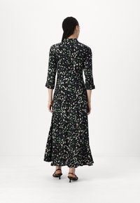 Robe maxi noire avec un motif de feuilles vertes et roses, manches 3/4, taille cintrée et ourlet fluide, assortie avec des sandales noires.