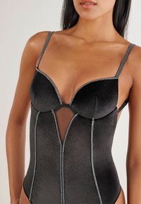 Sort bodysuit med strukturerede skåle, gennemsigtig mesh-panel og sølvfarvede ornamenter langs sømmene og stropperne. Glat tekstur.