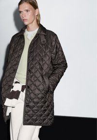 Hnědý prošívaný kabát s límcem, se skládajícími černými knoflíky a dvěma bočními kapsami, nošený přes světle zelený svetr a bílé kalhoty.