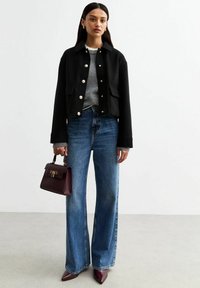 Svart kort jacka med stora knappar, grå tröja, högmidjade blå vida jeans och vinröd handväska. Spetsiga lila klackar.