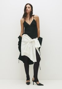 Sort satin camisole med blondekant, hvid skjorte bundet om taljen, sorte strømpebukser og spidse sorte hæle med dekorative detaljer.