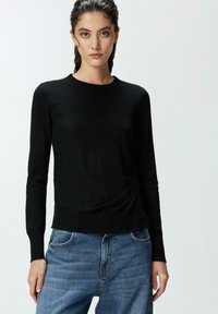 Top lungo a maniche nere in maglia con scollo rotondo, caratterizzato da una texture drappeggiata sul lato e un orlo a costine aderente. Il modello indossa jeans in denim blu.