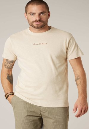 Homme aux cheveux courts et barbe portant un t-shirt beige avec le texte « Sun in the back » et un pantalon marron clair à motifs, mains détendues.