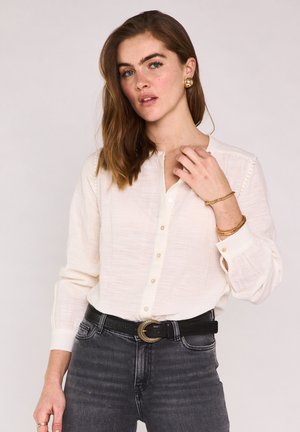 Jeune femme aux cheveux bruns ondulés portant un chemisier crème boutonné, un jean foncé, des bijoux dorés et une ceinture noire avec une boucle décorative.