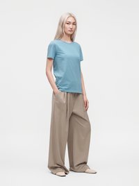 Jeune femme aux longs cheveux clairs, debout, portant une chemise bleue à manches courtes, un pantalon beige large et des chaussures claires sur fond blanc.
