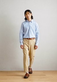 Chemise bleu clair à manches longues, chinos beiges, mocassins marron, debout sur un sol en bois devant un mur uni, cheveux de longueur moyenne et foncés.