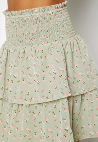 Jupe mini florale en vert clair avec une taille froncée. Présente des volants superposés et un motif de petites fleurs roses et de feuilles vertes.