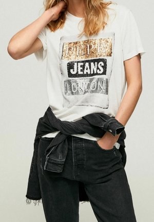 Osoba nosząca białą koszulkę z tekstem z cekinów w kolorze złotym, czarnym i srebrnym "PEPE JEANS LONDON,", czarne dżinsy oraz czarną kurtkę zawiązaną w talii.