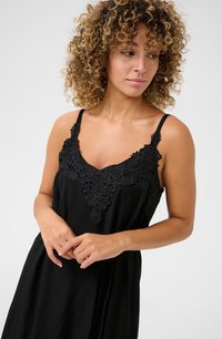 Vêtement noir avec des détails en dentelle au niveau du col, doté de fines bretelles réglables et d'une coupe fluide. Tissu doux et texturé.