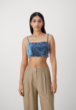 Avskåret blå denimtop med spaghettistropper, frynsete kant og subtile pyntedetaljer. Kombinert med lys beige, løstsittende bukser.