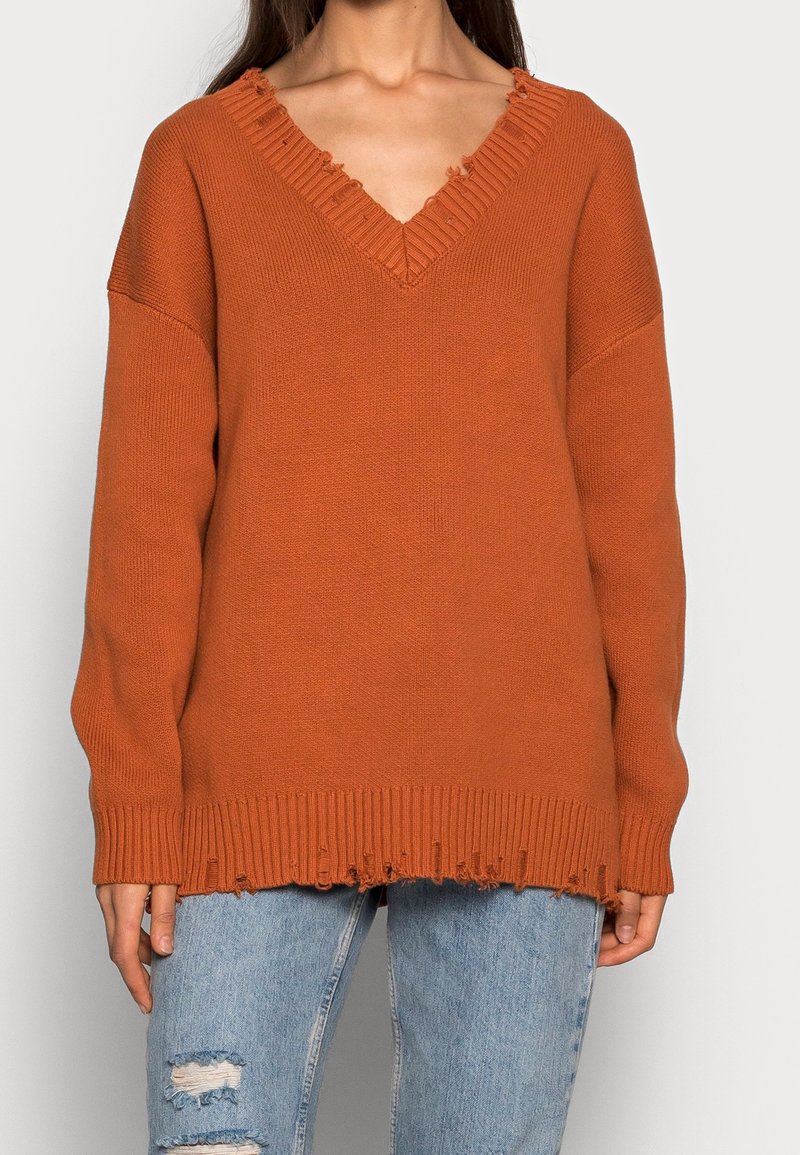 Stickad tröja i rostfärg med V-ringning, fransiga kanter och ribbad nederkant. Har långa ärmar och en ledig passform.