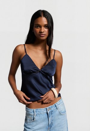 Giovane donna con capelli lunghi e scuri che indossa un top blu navy con spalline sottili e jeans di denim chiaro, in piedi davanti a uno sfondo semplice.