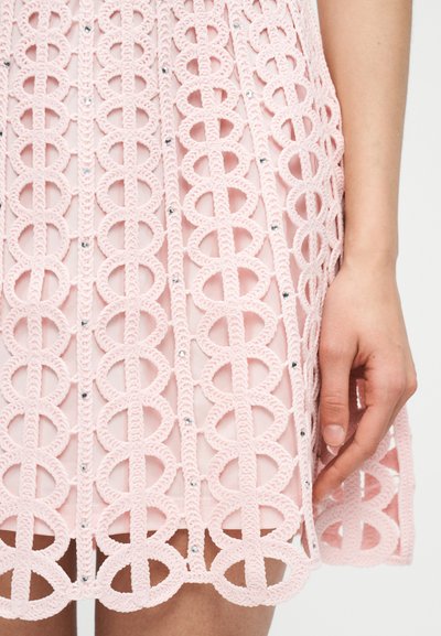 Κοντινό πλάνο ενός ροζ πλεκτού φούστας με κυκλικά σχέδια και μικρές στρας, φορεμένη από ένα άτομο με το δεξί χέρι χαλαρό στο πλάι.